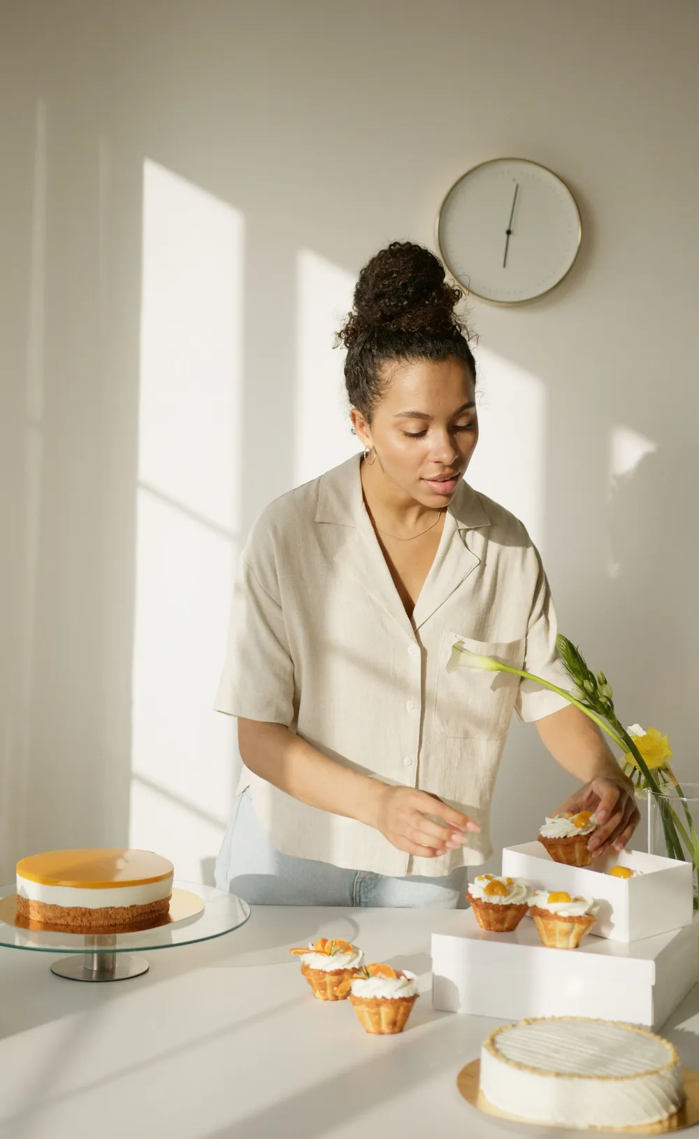 A baker in a sunlit kitchen boxing cupcakes beside finished cakes on a stand.