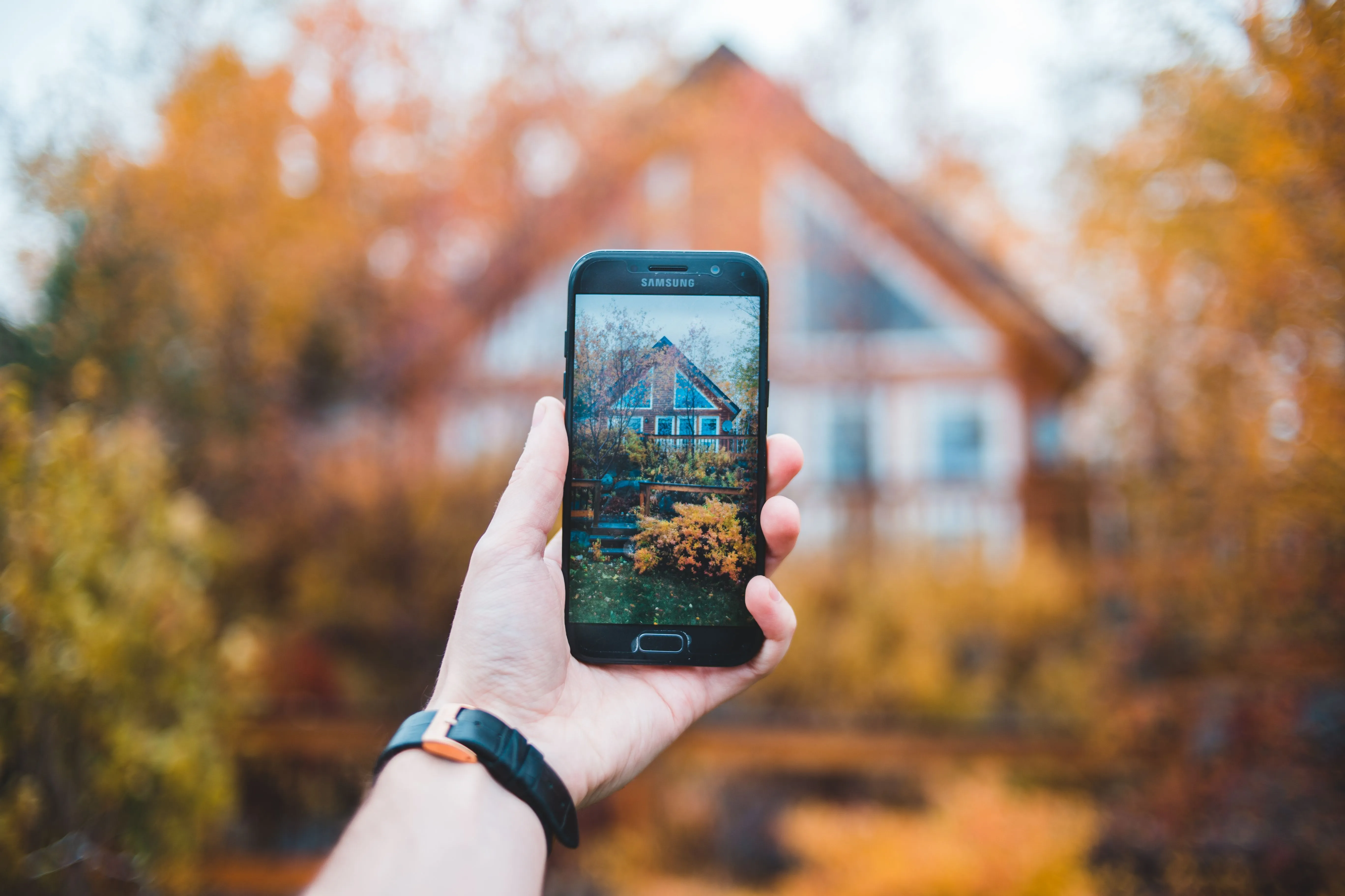 A hand holding a phone up to a gabled house in autumn, the finished photograph of that house filling the screen.