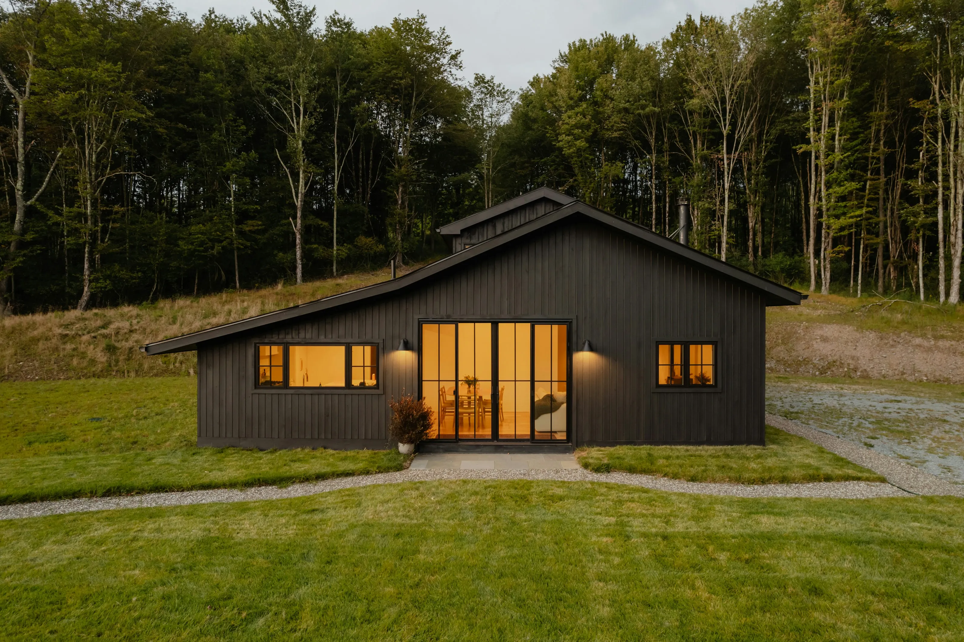A black board-and-batten house photographed head on at dusk, its windows and glazed doors lit from within against the treeline behind it.