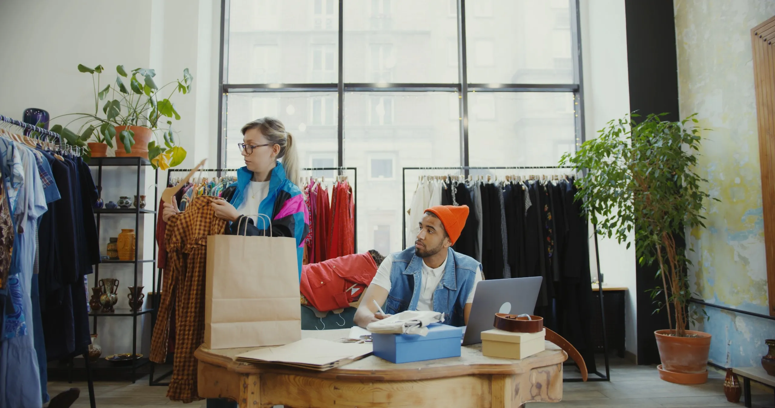 Two shopkeepers at a work table in a sunlit clothing store, sorting stock beside a laptop and a rail of garments.
