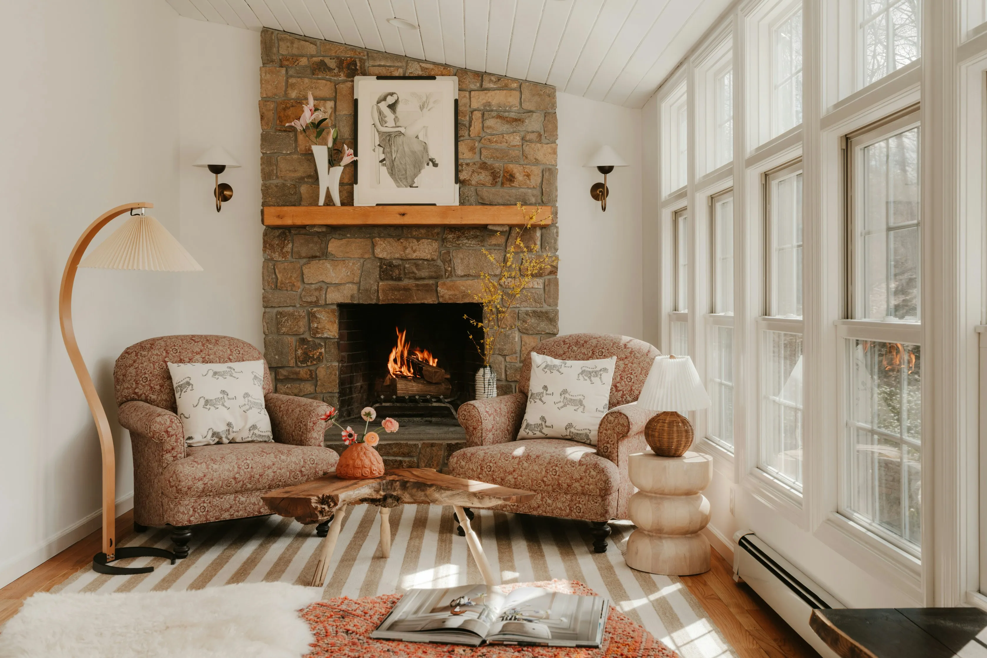 A stone chimney breast and timber mantel rising to a boarded ceiling, flanked by tall windows and a lit fire.