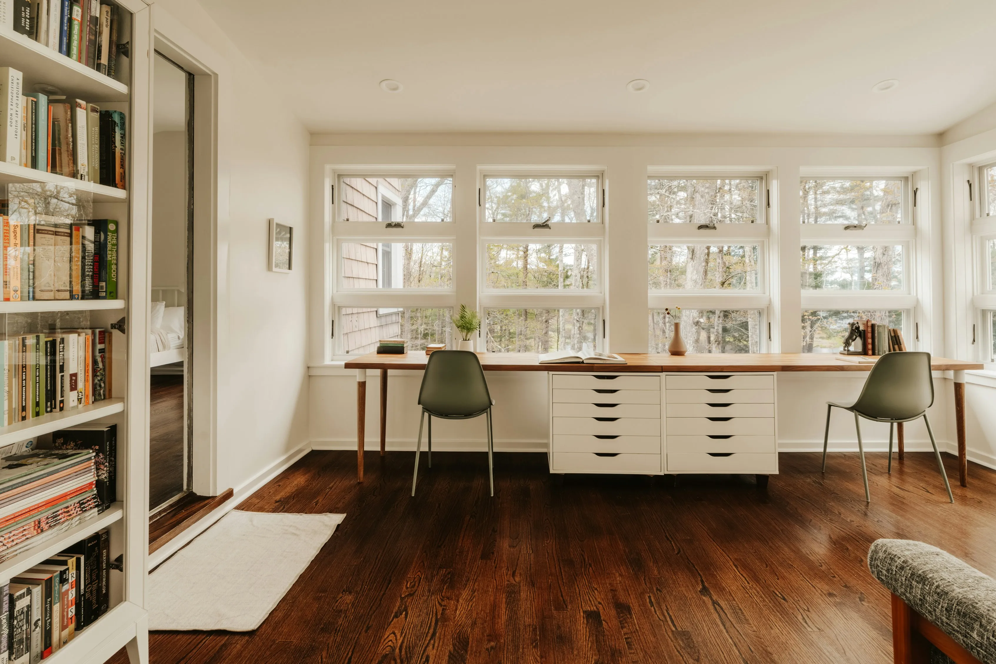 A study with a long timber desk running beneath a bank of windows, dark wood floors, and a full bookcase in the foreground.