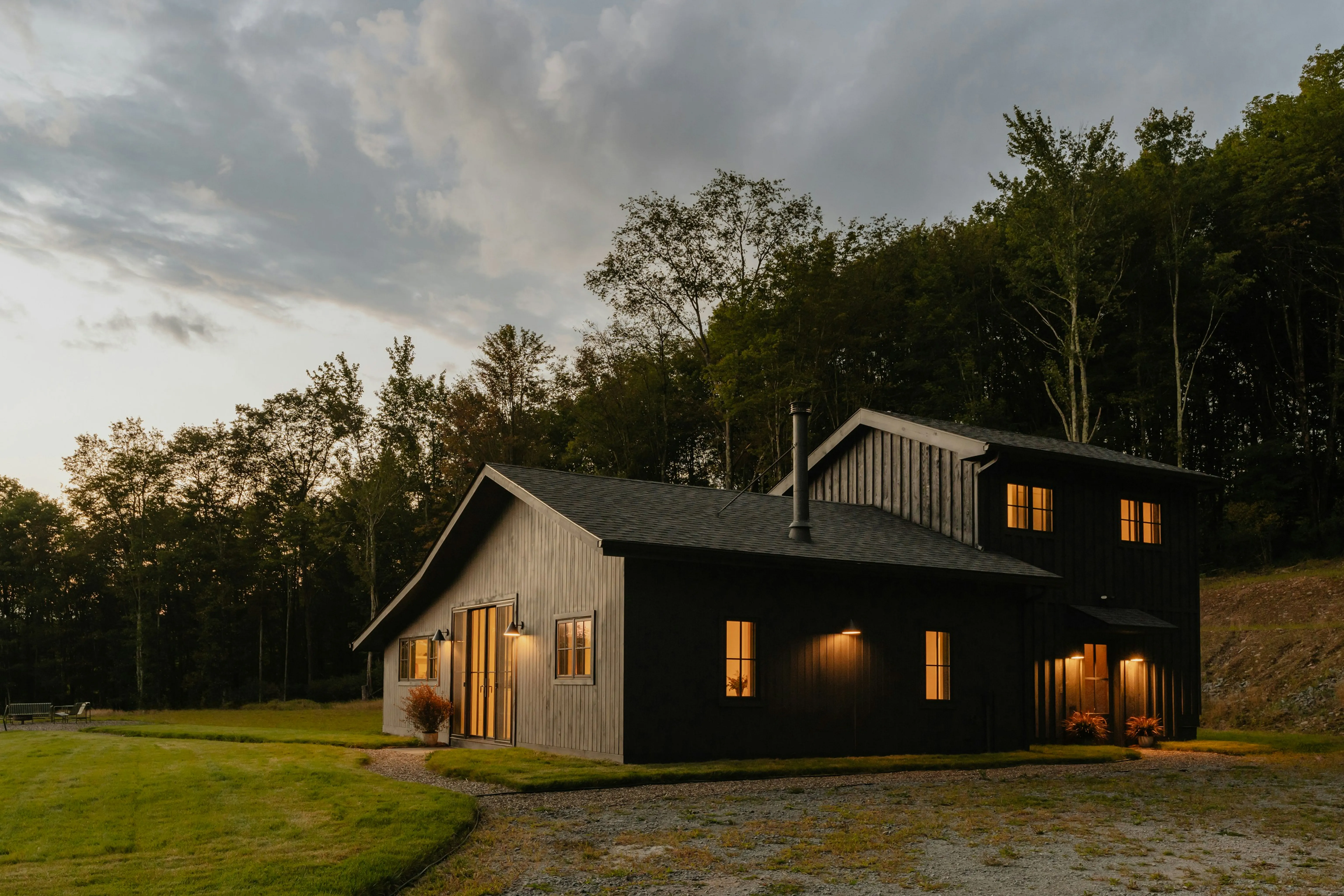 A modern barn-style home photographed at dusk, its windows lit from within, set against a treeline above a gravel drive.